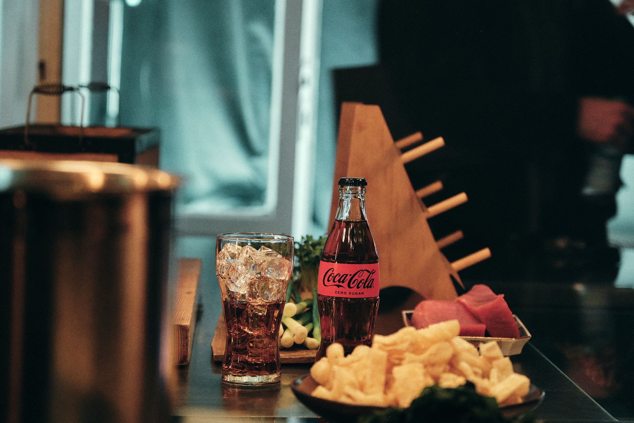 Eine Coca-Cola Zero Sugar Flasche steht neben einem mit Eiswürfeln gefüllten Glas auf einem Restauranttisch; im Vordergrund ist ein Teller mit Speisen zu sehen.
