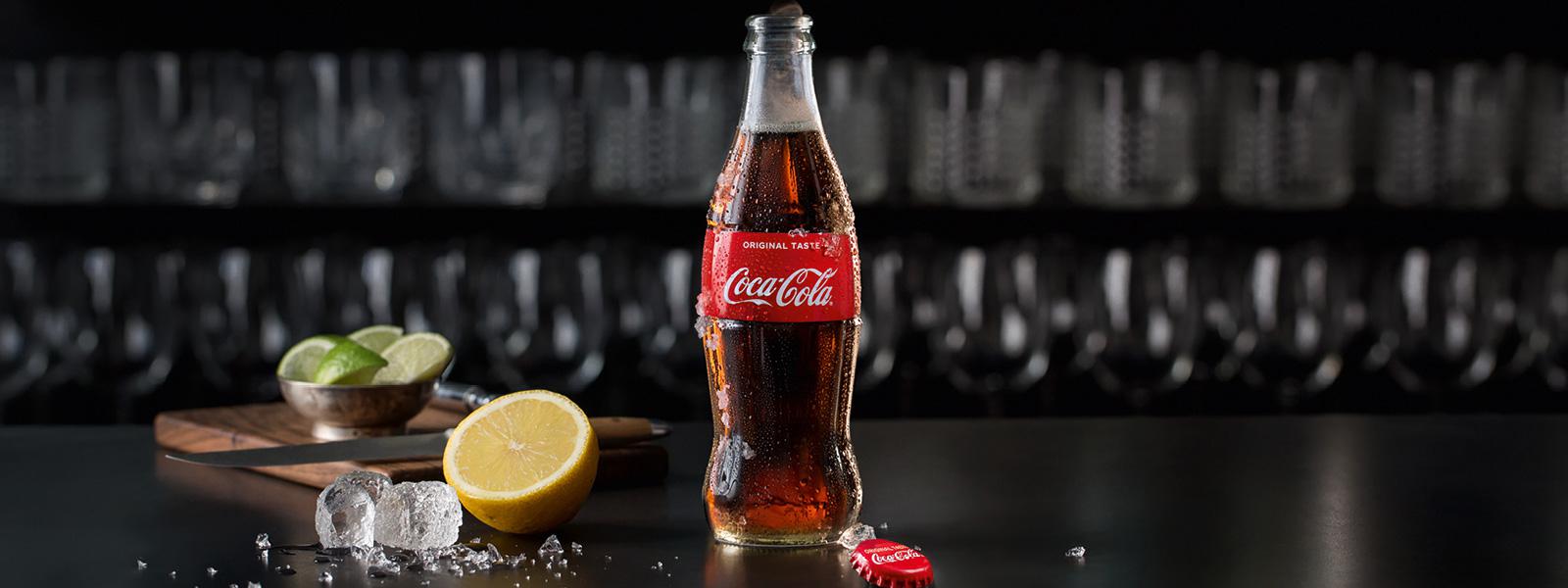 En glasflaske med Coca-Cola Original smag står på bordet sammen med en citron
