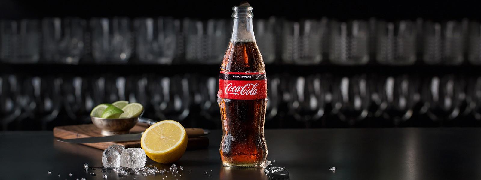 En glasflaske med Coca-Cola Original smag står på bordet sammen med en citron