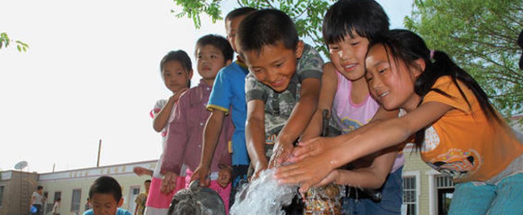 En række med smilende asiatiske børn står med hænderne samlet i en opadgående vandstråle
