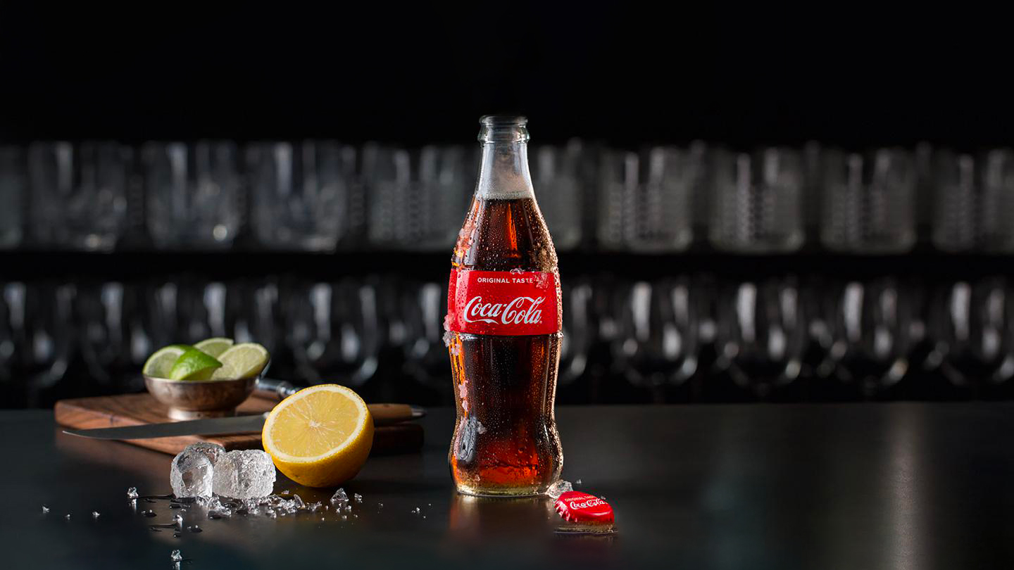 Åben Coca-Cola-glasflaske med synlige dråber står på et bord ved siden af citron, lemon, isterning og skærebræt med kniv