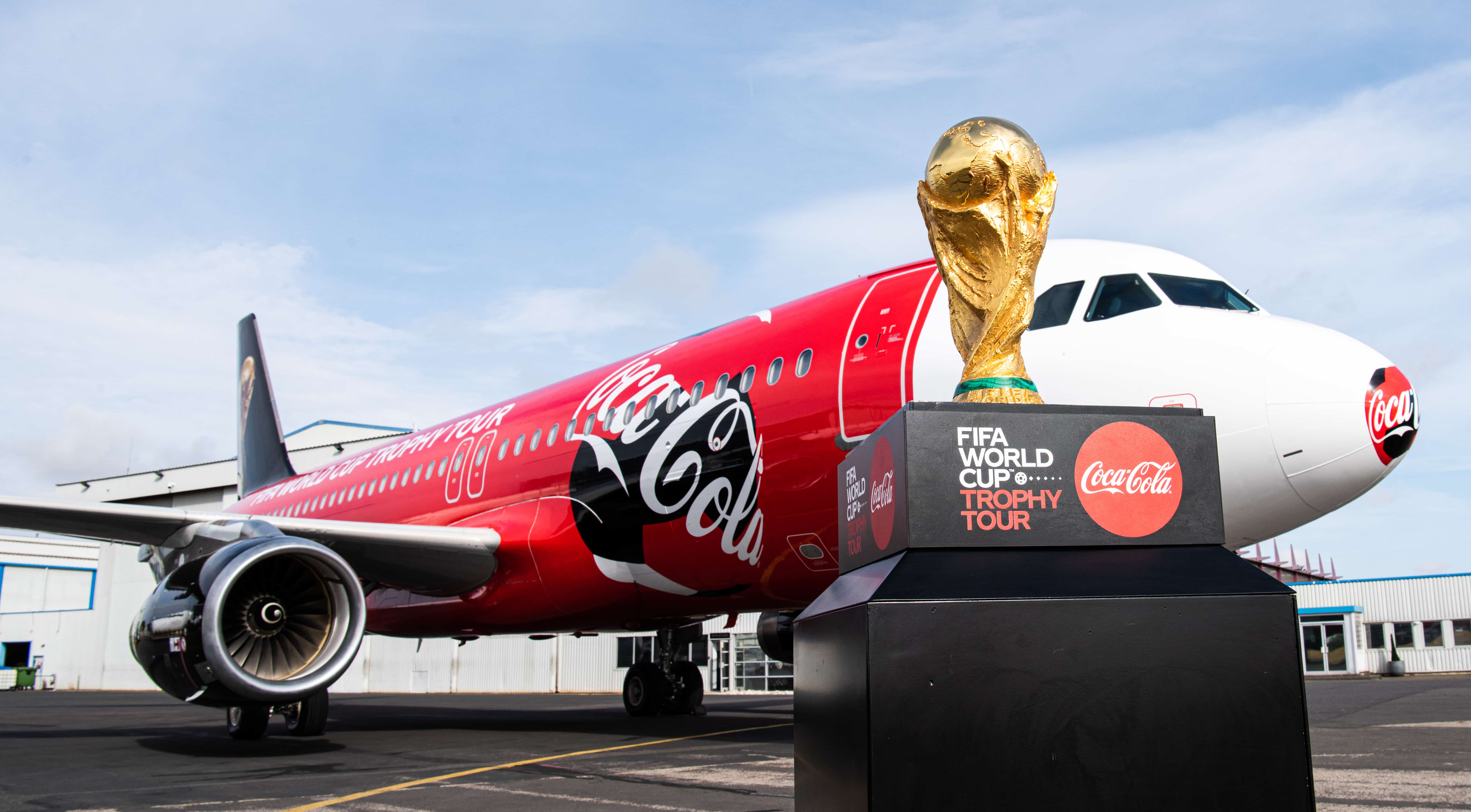 Fodbold-VM-trofæ står på et lille podie med Coca-Cola-branding og med et fly dekoreret i Coca-Cola-farver i baggrunden