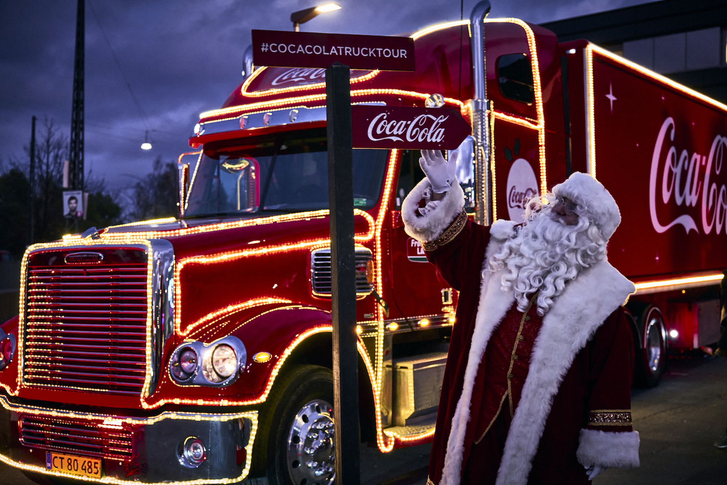 Julemanden står foran oplyst rød Coca-Cola-lastbil og skilt med vejvisning