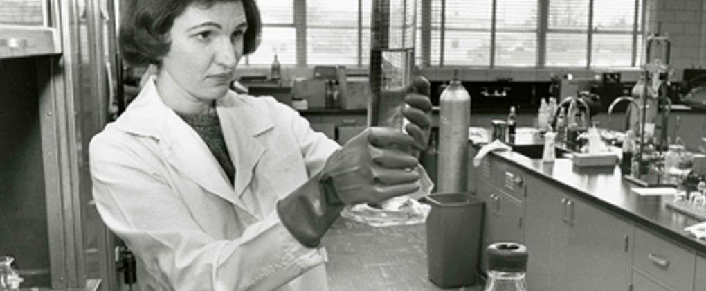Sort/hvid-billede af kvinde i kittel i laboratorie med kolbe i hænderne