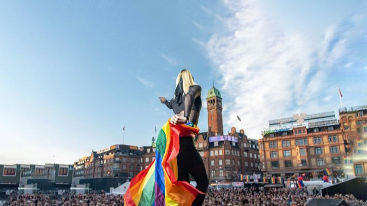 Lyshåret kvinde med regnbuefarvet flag står med ryggen til på en scene i København foran publikum
