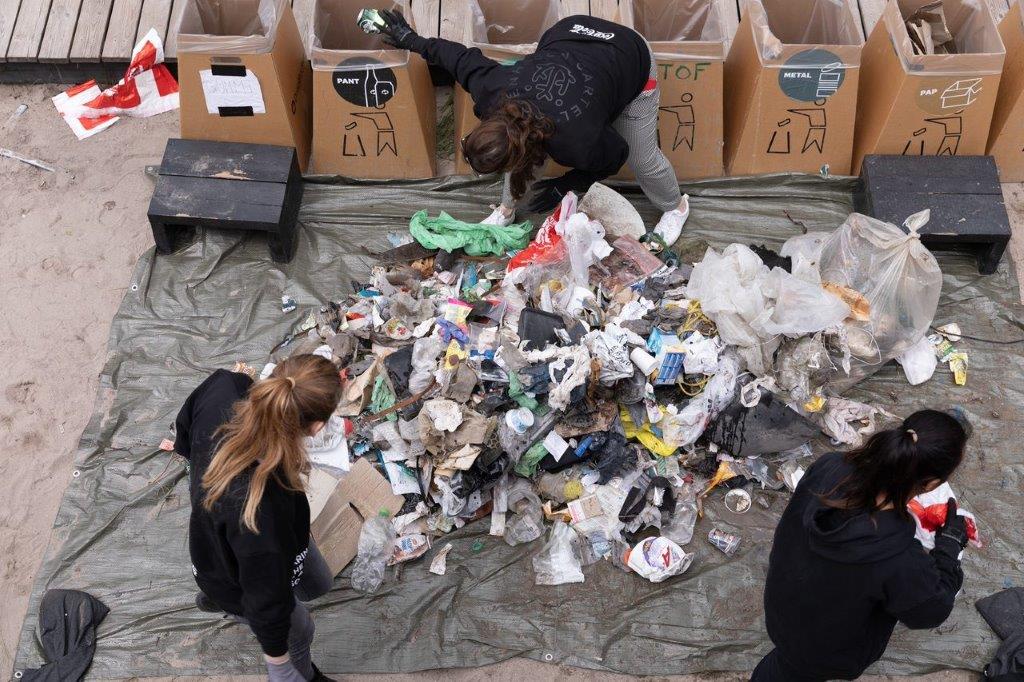 Tre kvinder sorterer en affaldsbunke på en presenning i brune affaldskasser