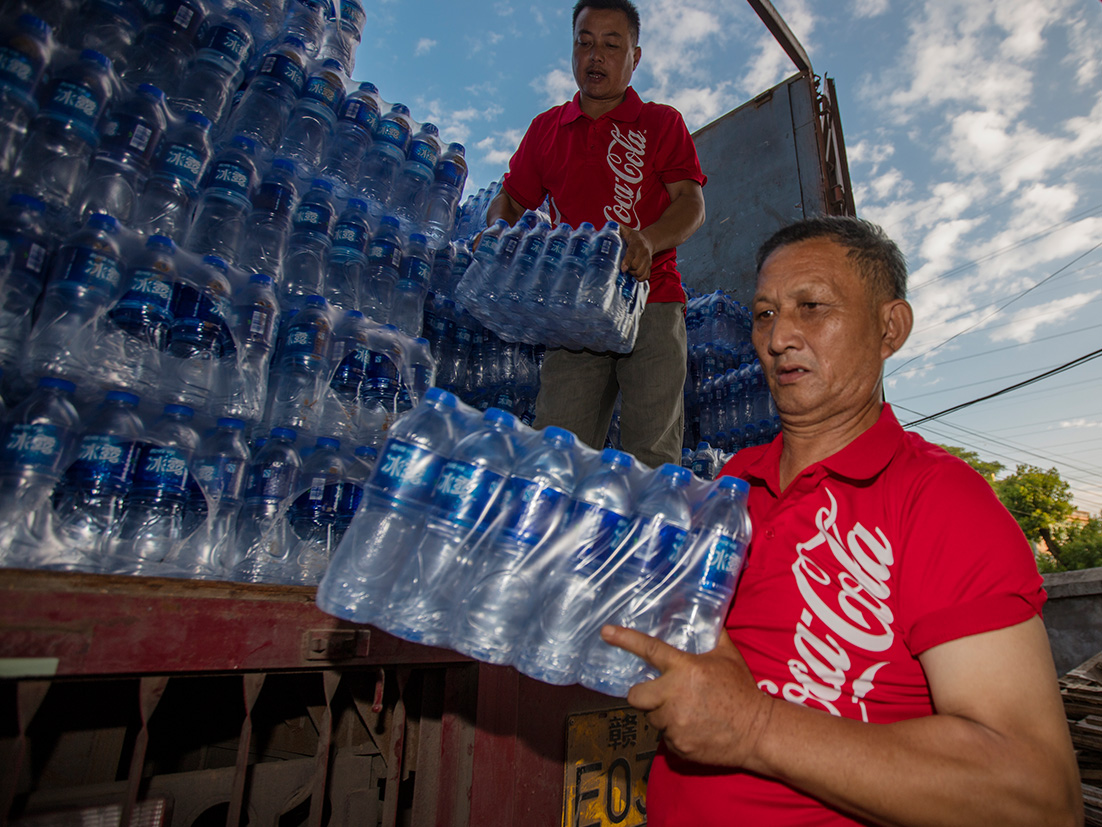 To mænd i røde Coca-Cola-trøjer løfter pakker med vand ned fra åbent lad på lastbil
