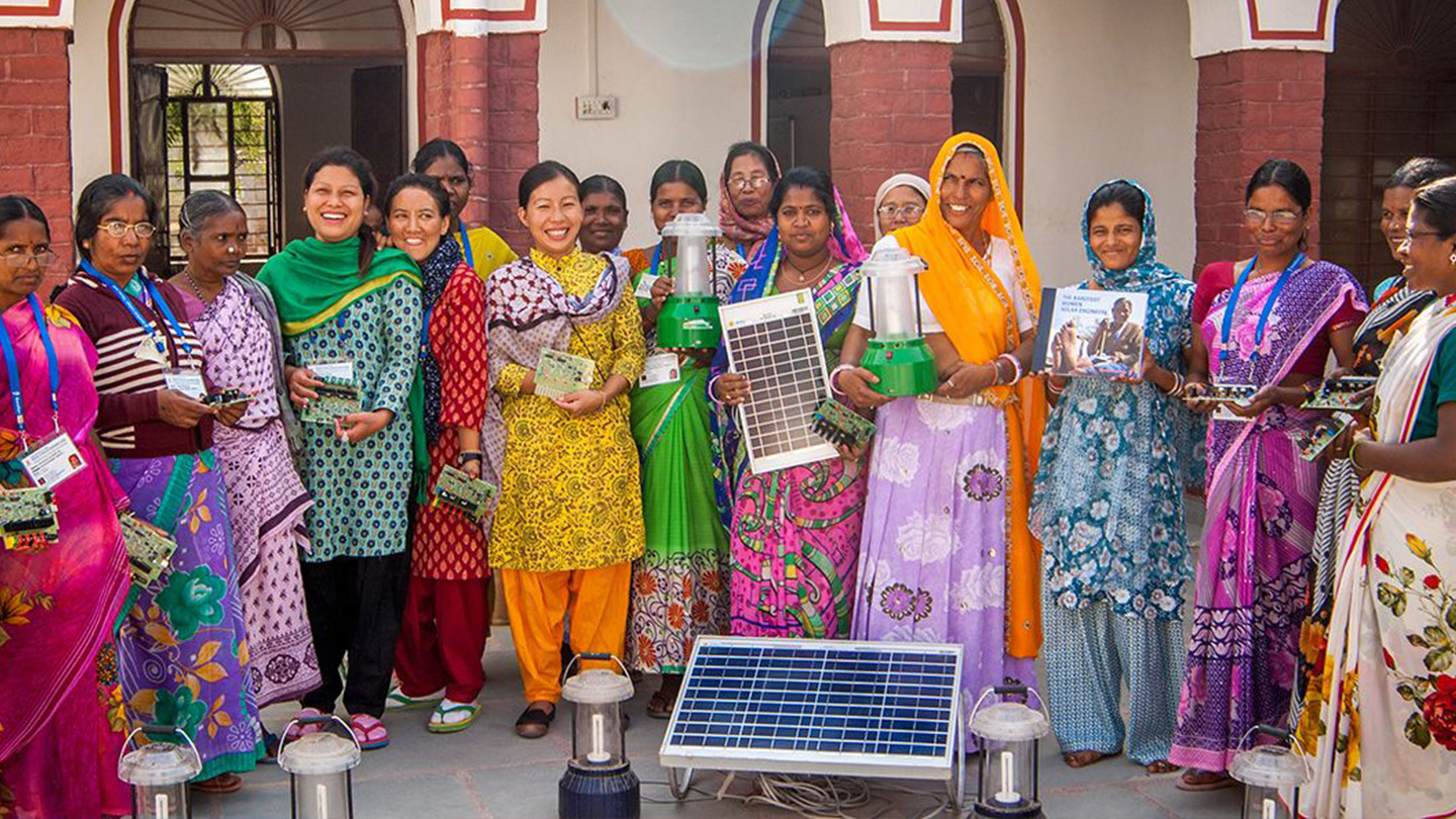 18 indiske kvinder i farverige kjoler står på en række foran en bygning med solpaneler i hænderne og foran dem
