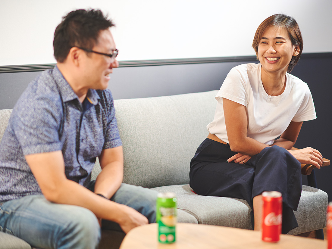 En smilende mand og kvinde sidder i en grå sofa med Coca-Cola-dåser i forggrunden på et sofabord