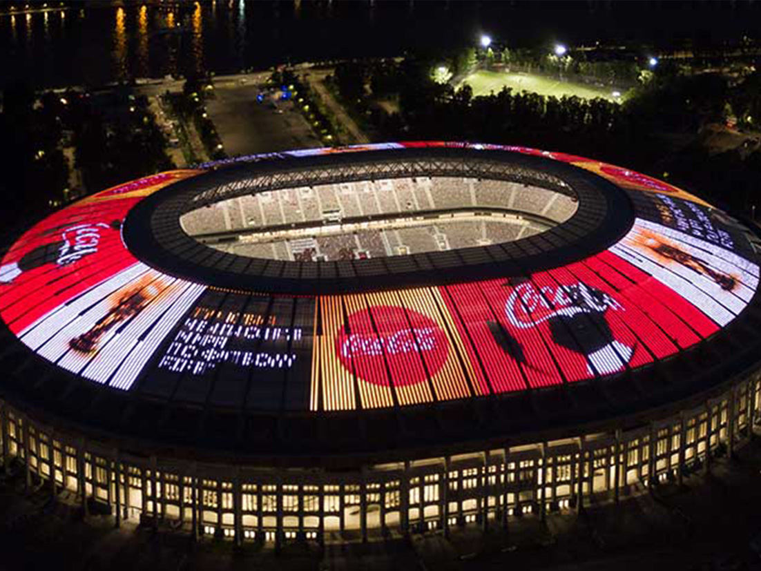 Sportsstadion set skråt oppefra i mørke med oplyste Coca-Cola-reklamer på stadiontag