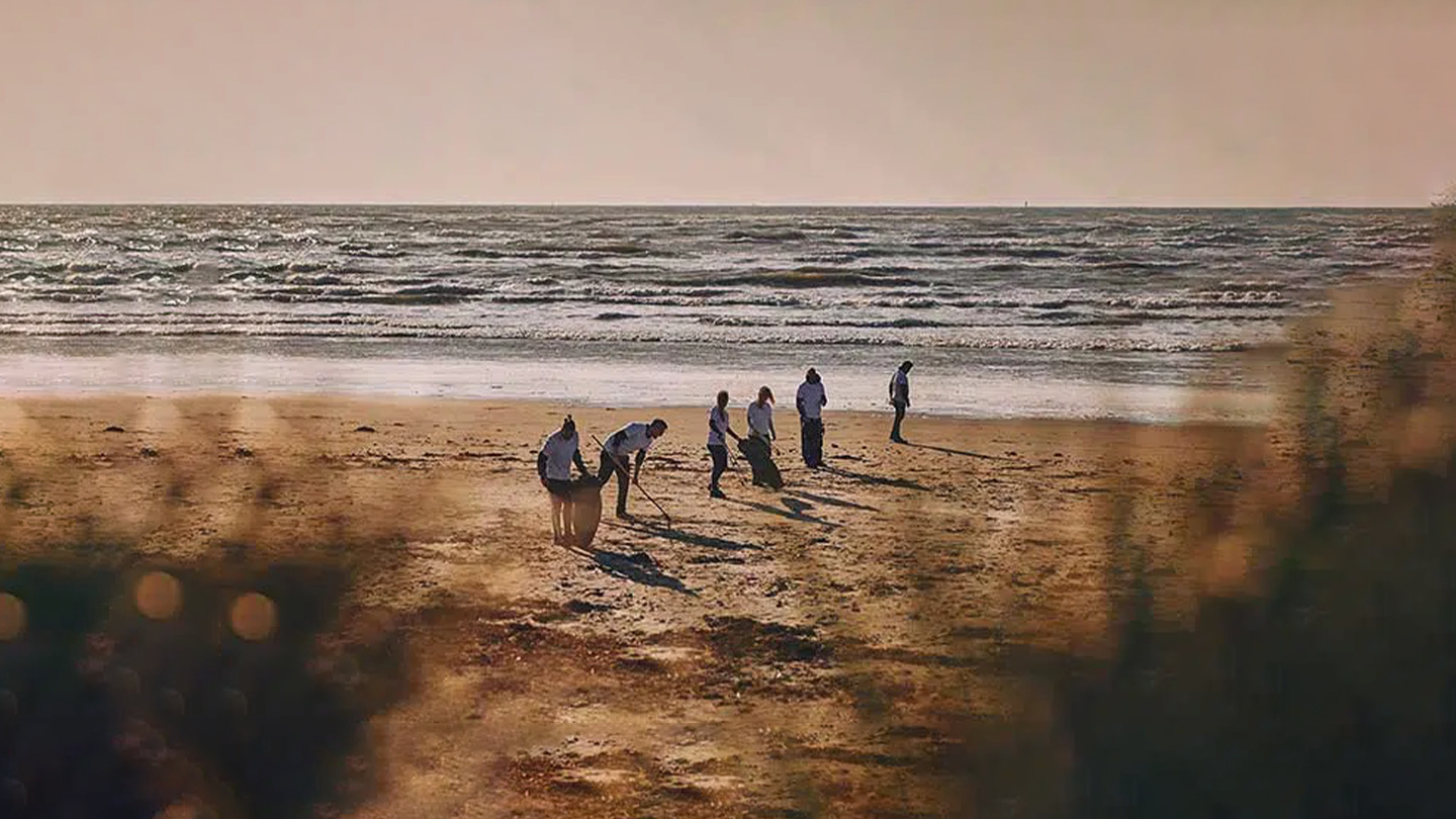 6 personer i hvide trøjer går i en række på en strand og indsamler affald i sorte affaldsposer