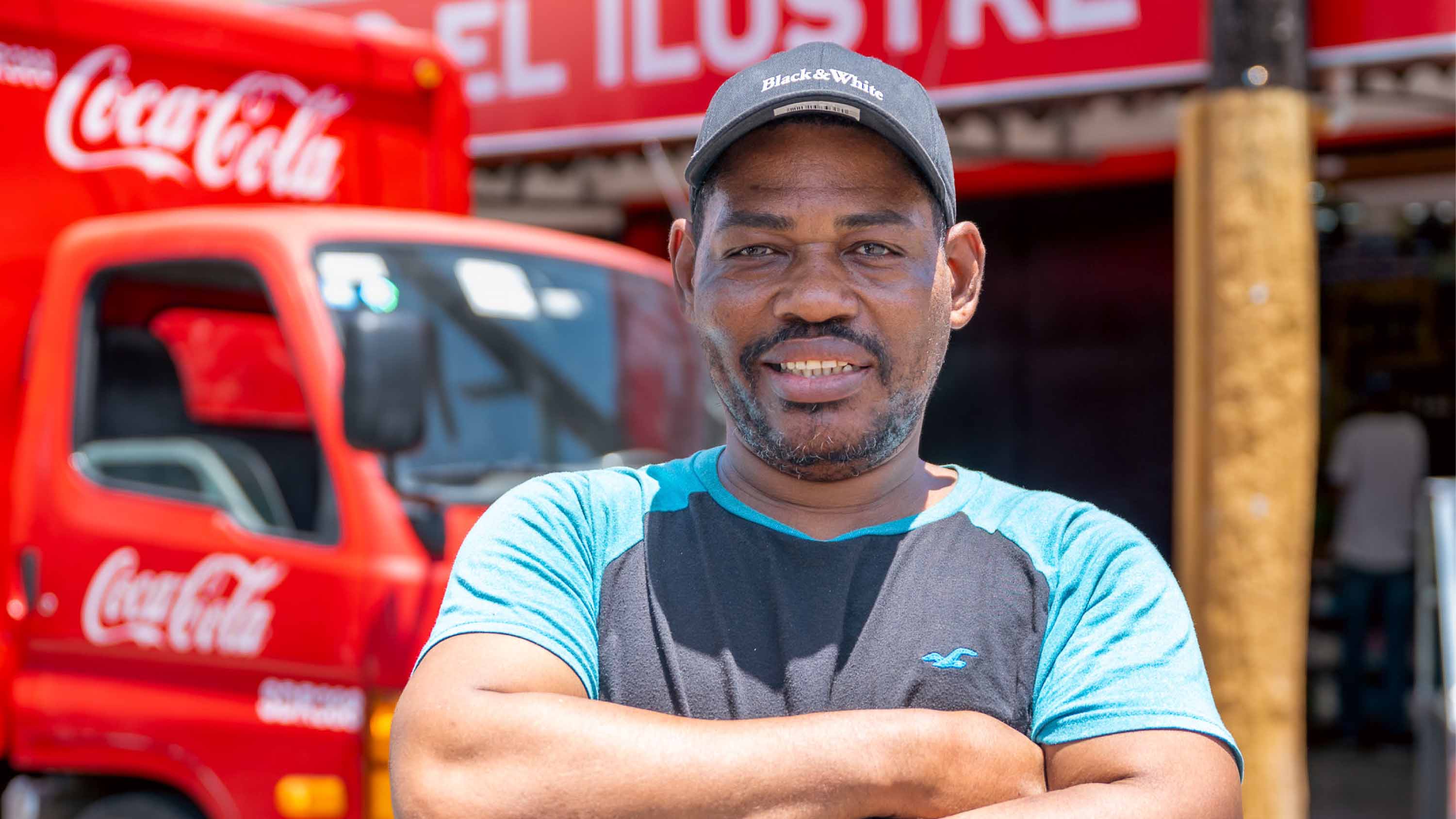Hombre sonriente de pie con los brazos cruzados frente a un camión de distribución de Coca-Cola.