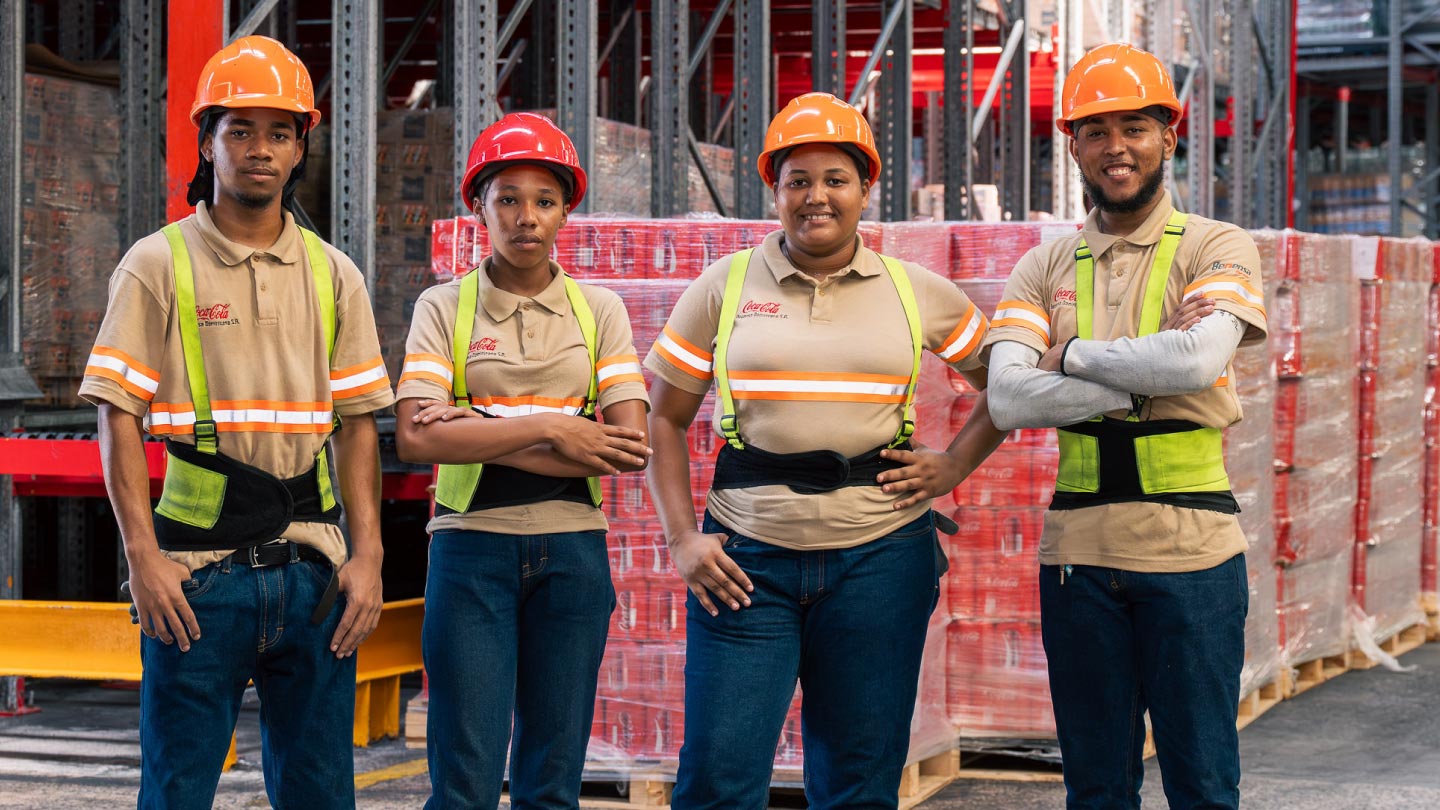 Cuatro empleados de Coca-Cola, con cascos de seguridad y uniformes de trabajo, posan en un almacén lleno de productos. 