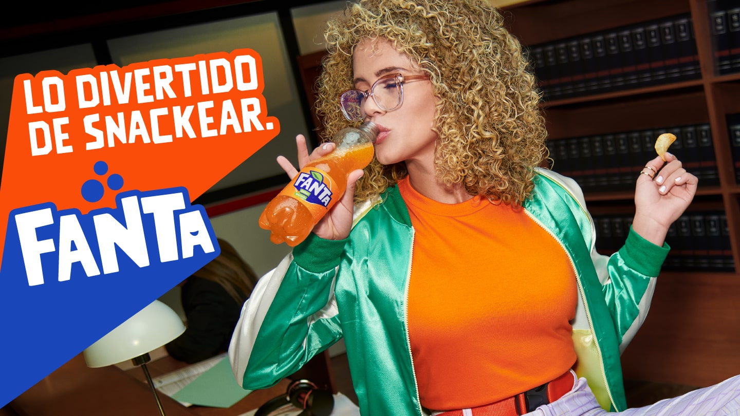 Mujer rubia con anteojos en una biblioteca vestida de manera divertida con una campera verde y una camisa naranja, bebiendo una Fanta refrescante y sosteniendo un gajo de naranja en su otra mano. A su derecha se observa el logo de Fanta y un texto que dice: 'Lo divertido de snackear'.