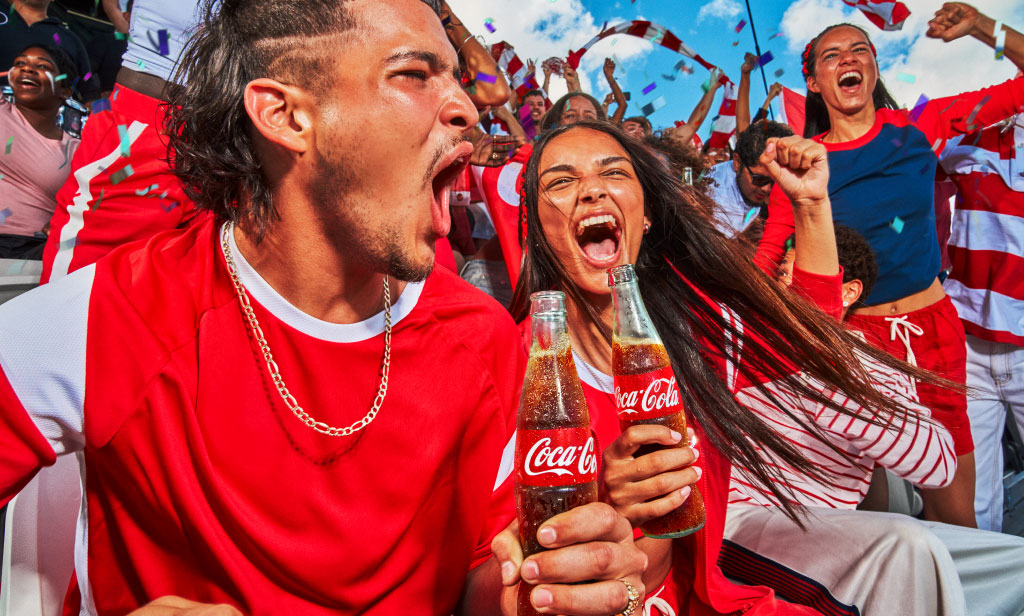 Grupo de aficionados vestidos de rojo celebrando en un ambiente festivo, con banderas y confeti, sosteniendo botellas de Coca‑Cola.