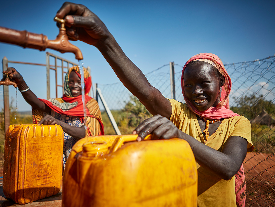 Kaksi hymyilevää afrikkalaista naista täyttää keltaisia kanistereita vesijohtovedellä.