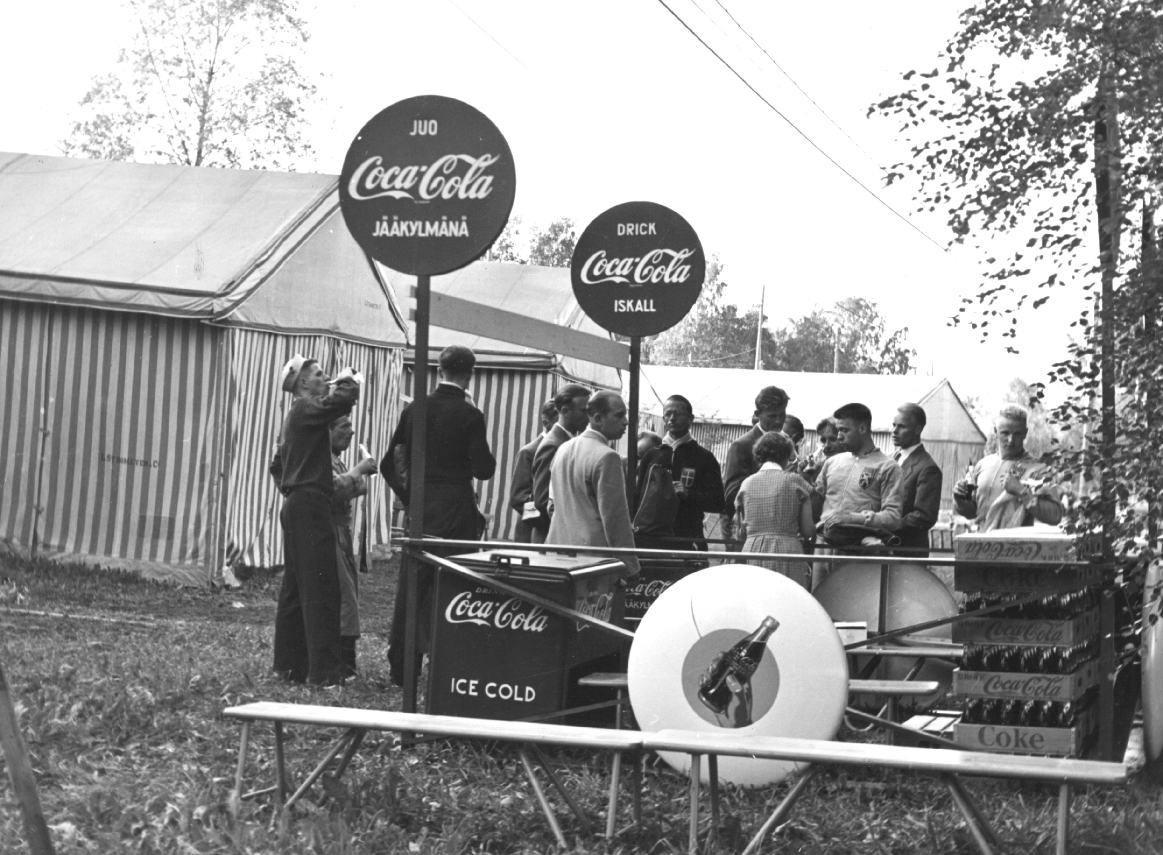 50-luvun Coca-Cola‑myyntikoju Helsingin kesäolympialaisissa. Retrokyltit,pullokorit ja ihmisiä juomassa Coca-Colaa.  