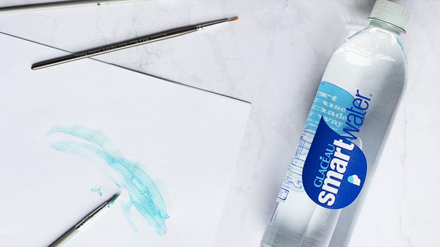 Une bouteille de Smart Water posée sur une table, à côté d'une feuille d'aquarelle et de pinceaux