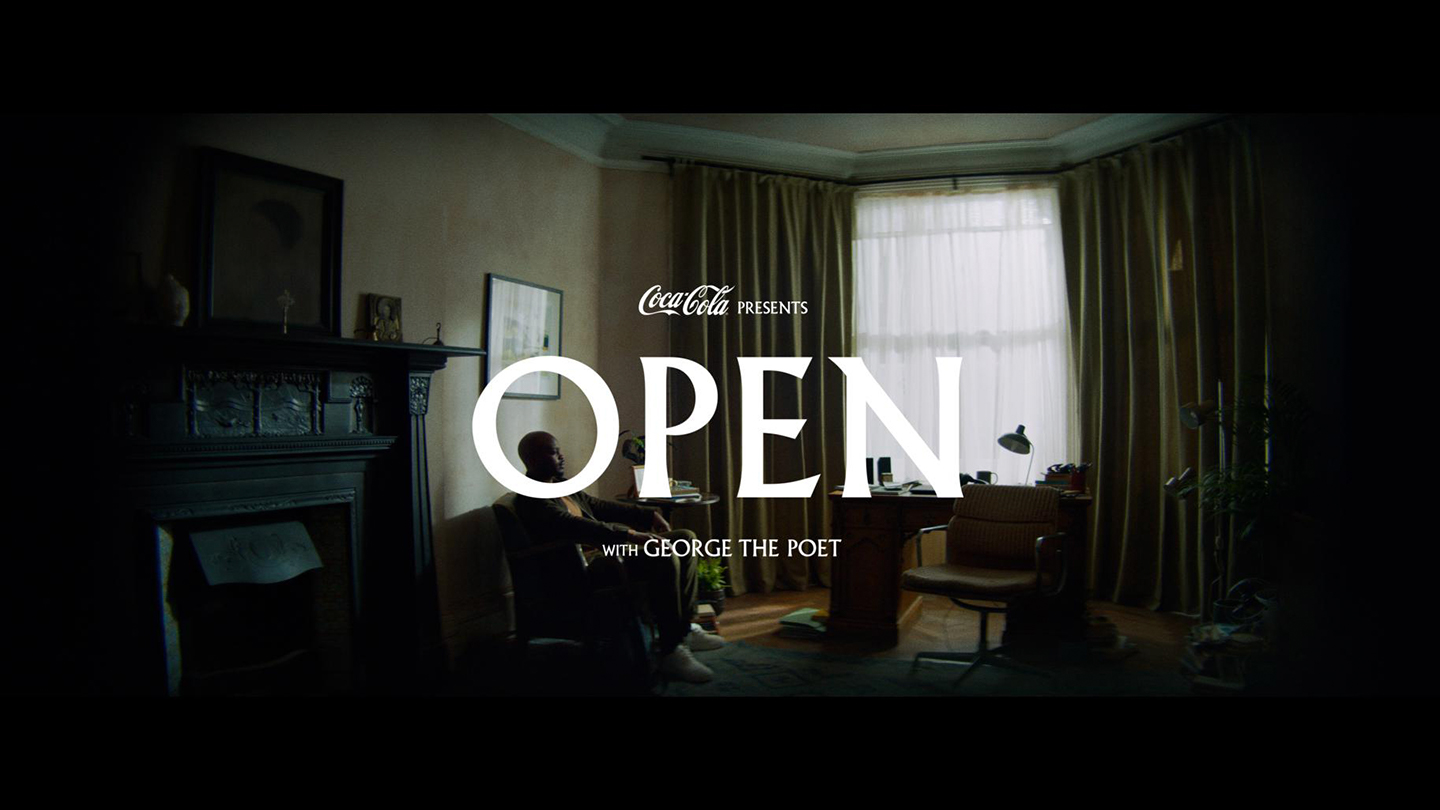 Interior of a house living room with low lighting with a young man sitting in a sofa; there is the following phrase displayed at the center of the image 'Coca-Cola presents OPEN with George The Poet