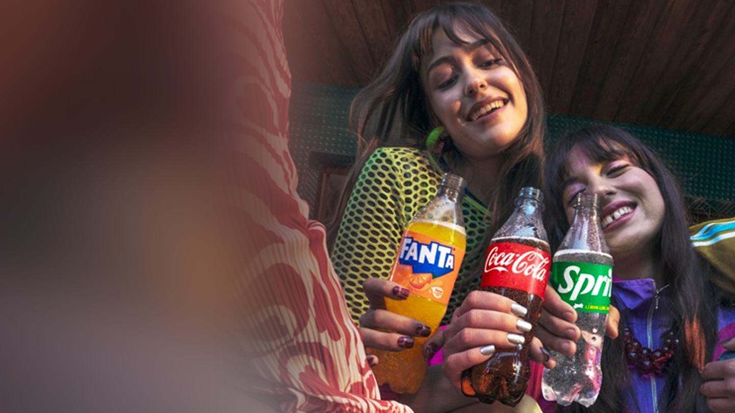 Personas sentadas juntas sosteniendo botellas de refrescos Fanta, Coca‑Cola y Sprite, en un ambiente relajado.