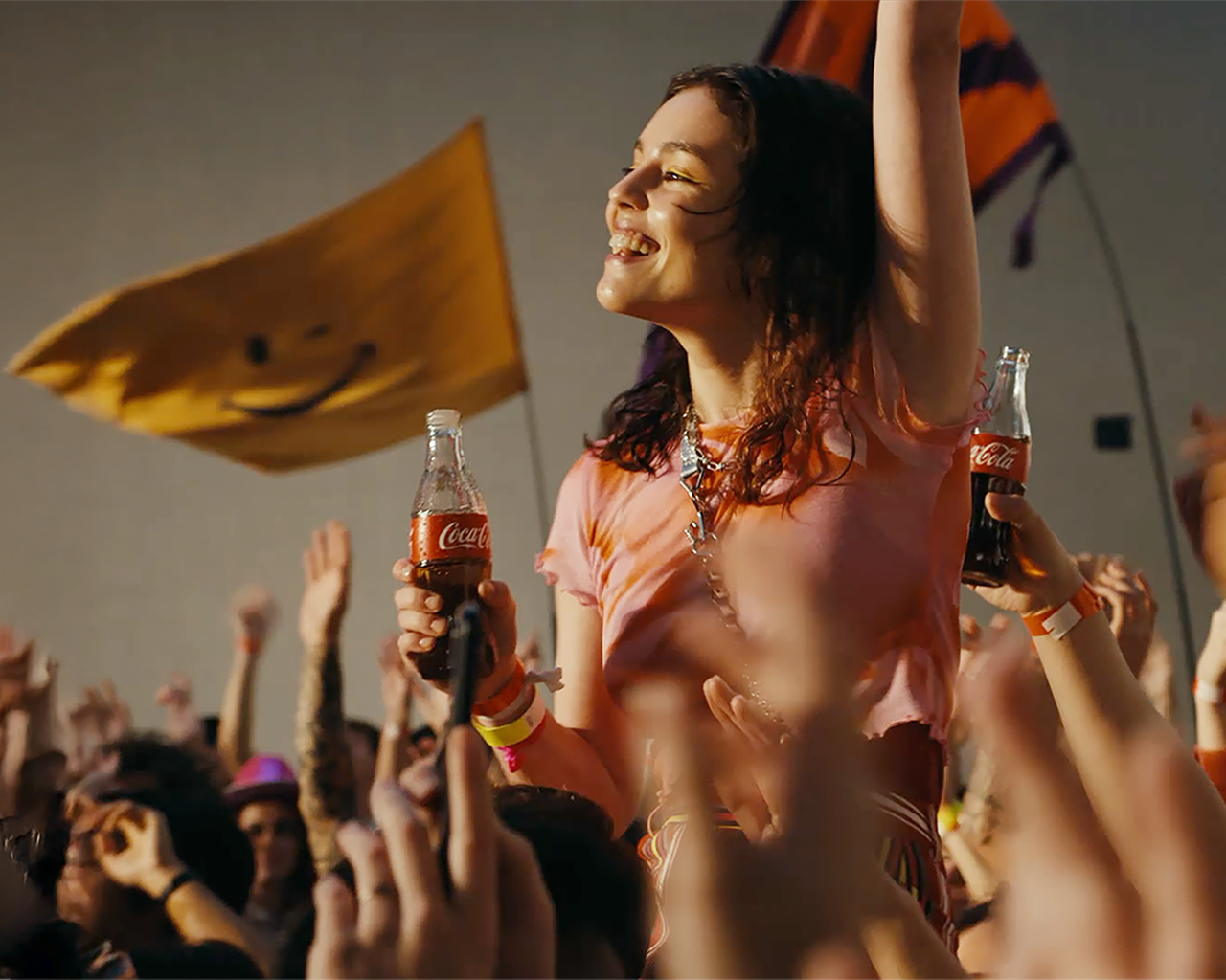 Persona celebrando en un concierto o festival al aire libre, sosteniendo una botella de Coca‑Cola, rodeada de una multitud con banderas y brazos levantados