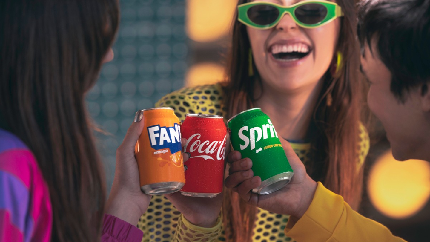 Several people clink cans of Fanta, Coca-Cola, and Sprite together.