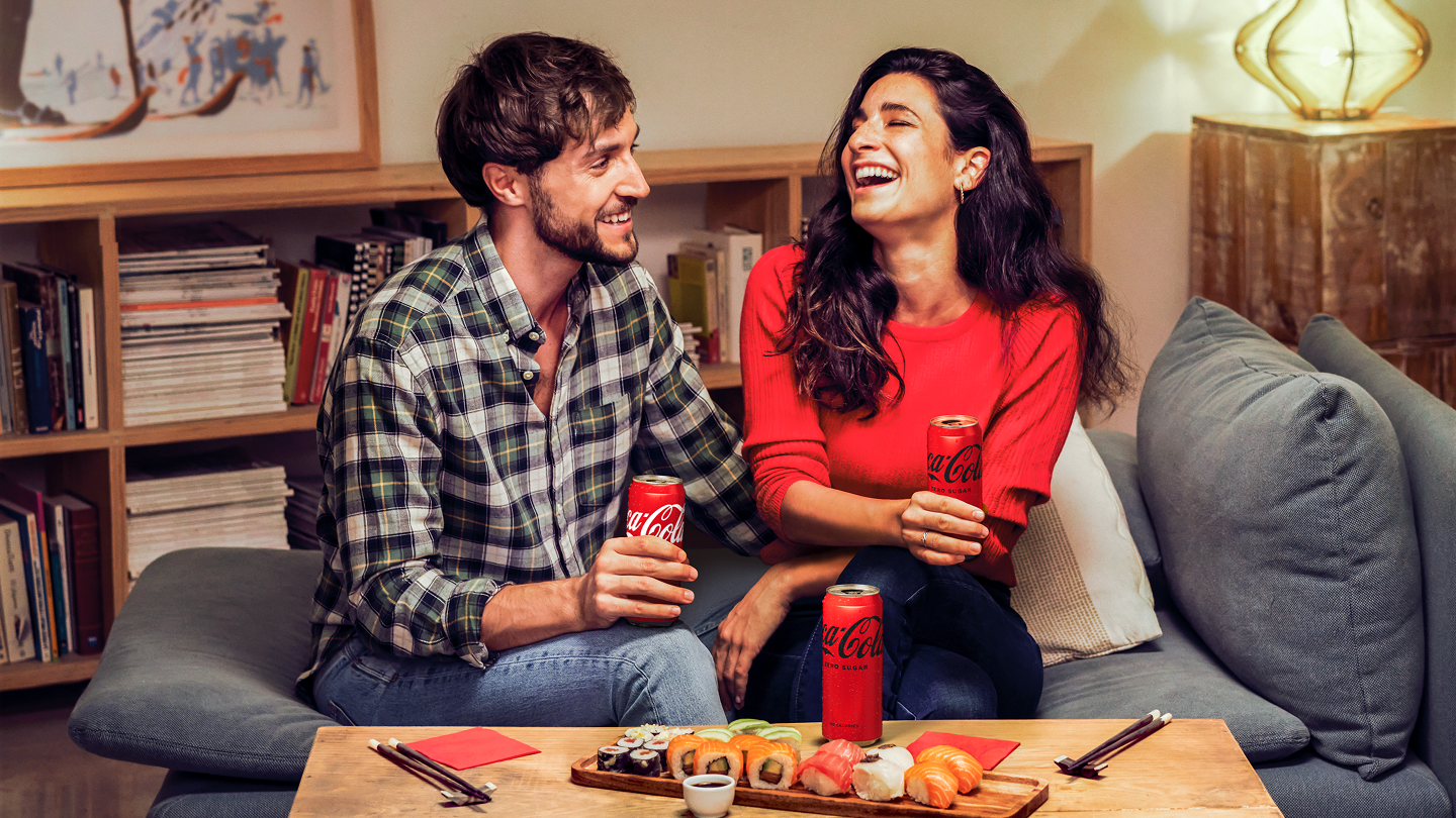 A man and woman, each holding a can of Coke, sit on a safa, laughing.