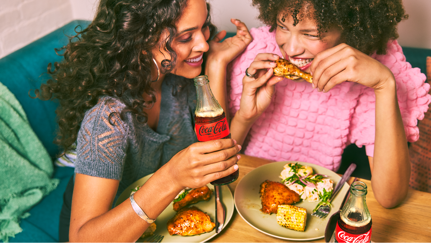 A couple of women sit around a table, eating chicken wings. They look like they are having fun. One woman raises her Coca-Cola Zero Sugar bottle.