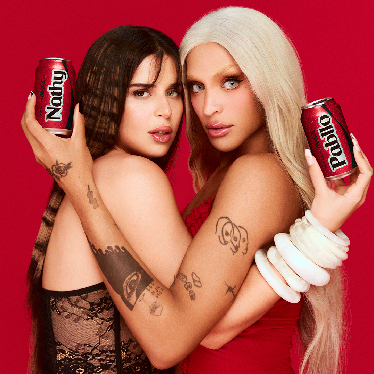 Nathy Peluso y Pabllo Vittar sostienen dos latas de Coca-Cola personalizadas con sus nombres, sobre un fondo rojo.