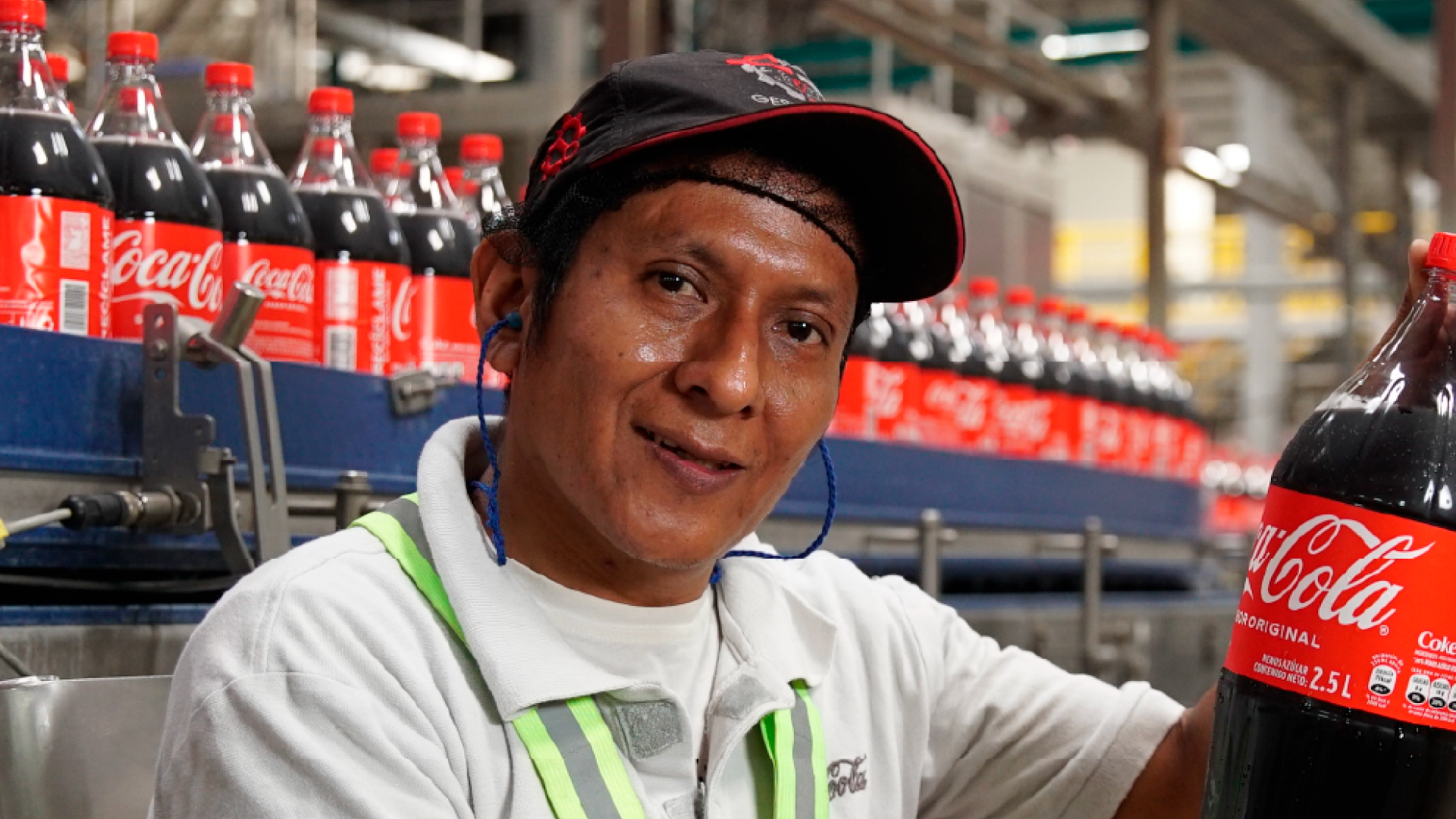 Hombre mirando a camara sonriente trabajando en una fabrica de Coca-Cola con una botella en la mano.