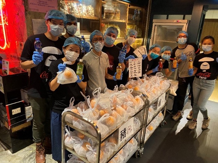 Group of people in an interior space behind a small stand with foods in plastic bags