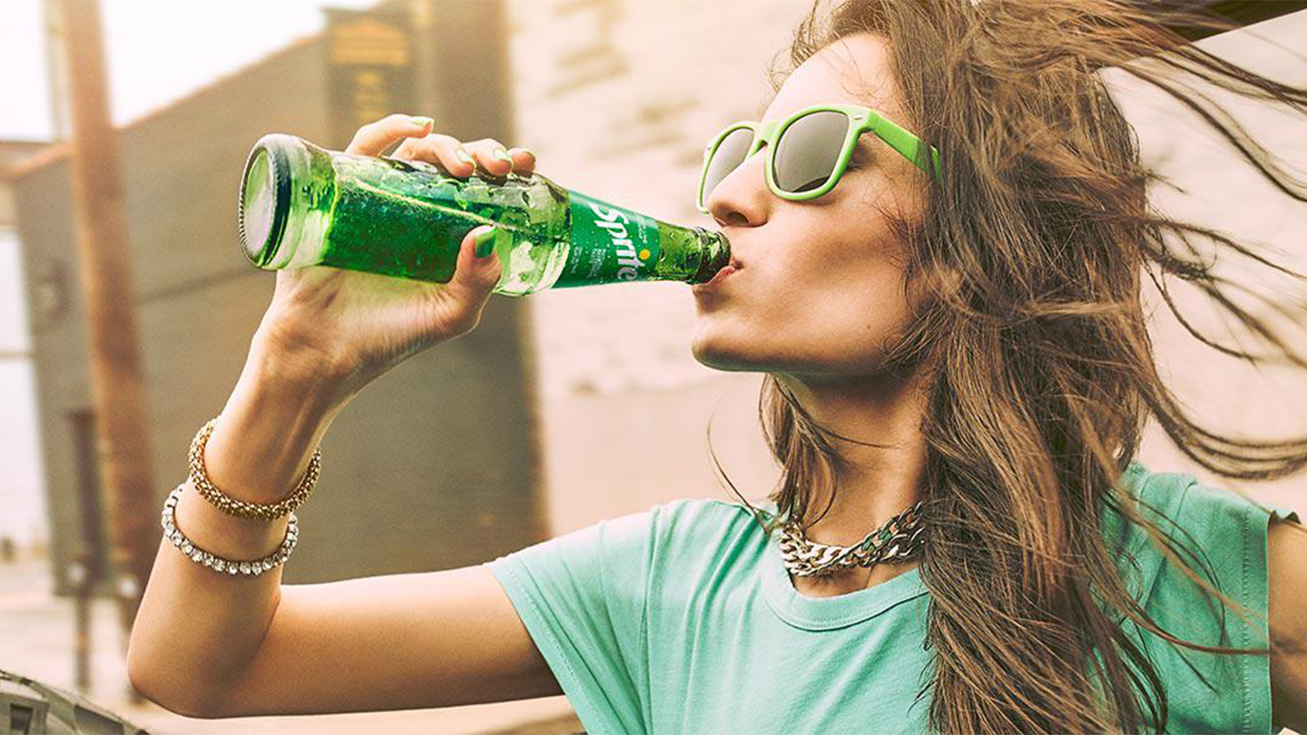 Djevojka u zelenoj majici i zelenim naočarima pije Sprite iz bočice