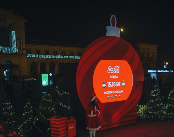 Coca‑Colina božićna instalacija na otvorenom s velikim LED ekranom koji prikazuje iznos donacije u sklopu humanitarne akcije, okružena blagdanskom dekoracijom i božićnim drvcima.