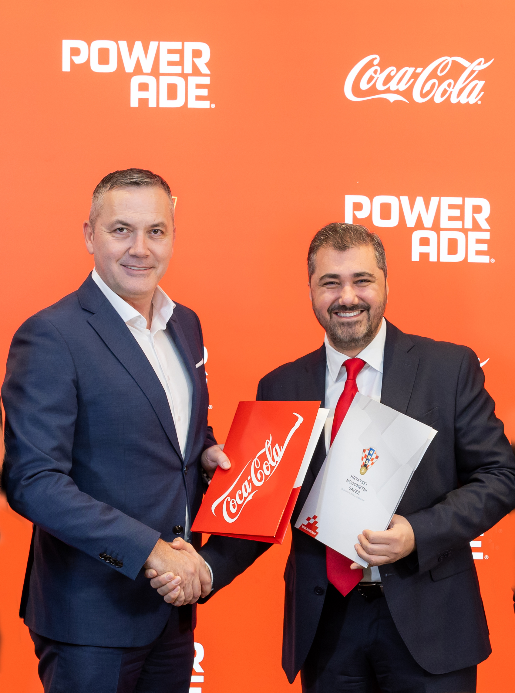 Representatives of the Croatian Football Federation and Coca‑Cola pose together holding branded folders in front of a Powerade and Coca‑Cola backdrop during the partnership signing event.
