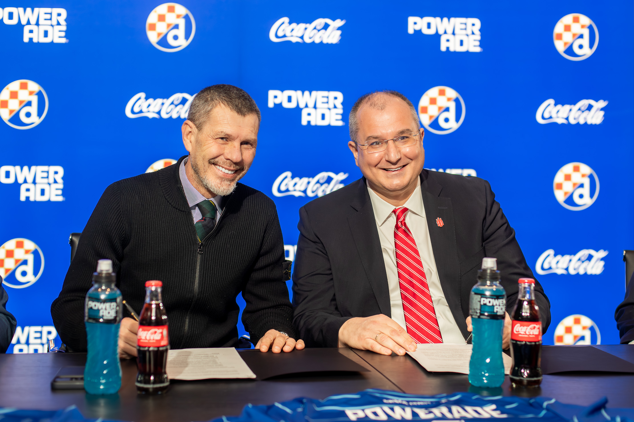 Predstavnici GNK Dinama i Coca‑Cole sjede za stolom tijekom potpisivanja sponzorskog ugovora, uz izložene proizvode Powerade i Coca‑Cola ispred brendirane pozadine.