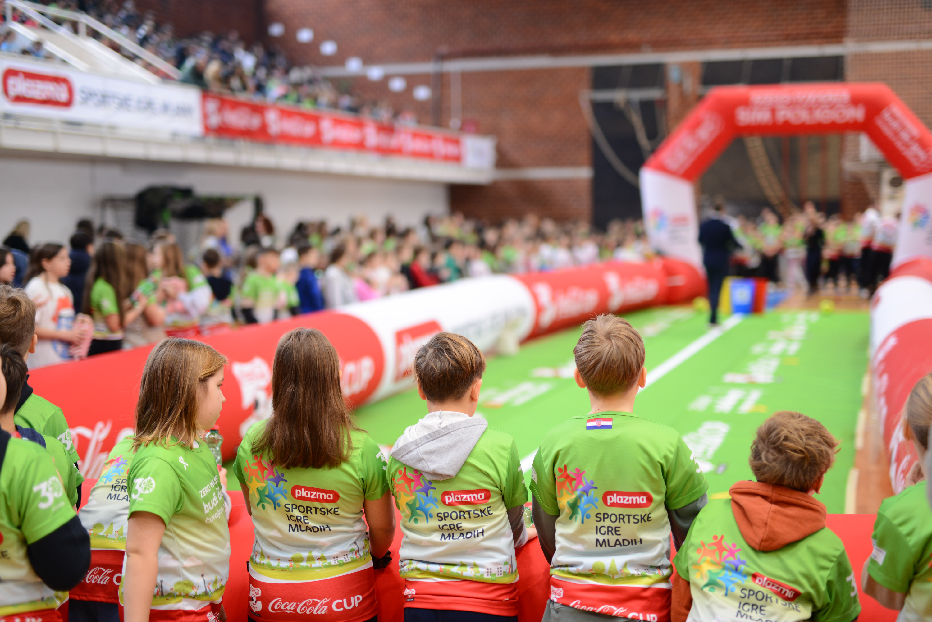 Djeca u sportskim dresovima stoje uz rub terena i prate događanja na nogometnom poligonu tijekom Coca‑Cola Cupa u sklopu Sportskih igara mladih.
