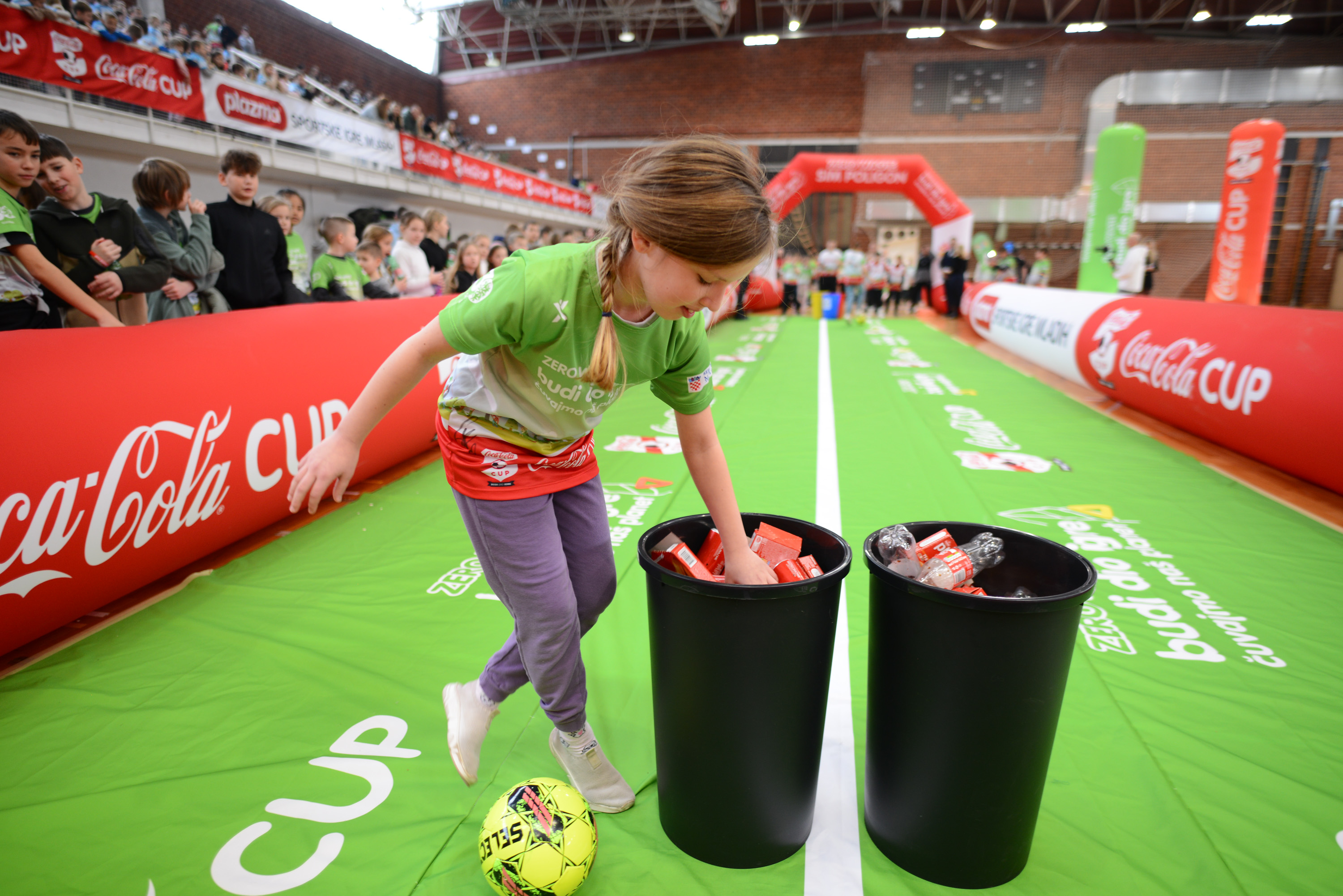 Dijete sudjeluje u edukativnoj Zero Waste aktivnosti tijekom Coca‑Cola Cupa, odlažući ambalažu u označene spremnike na sportskom poligonu.