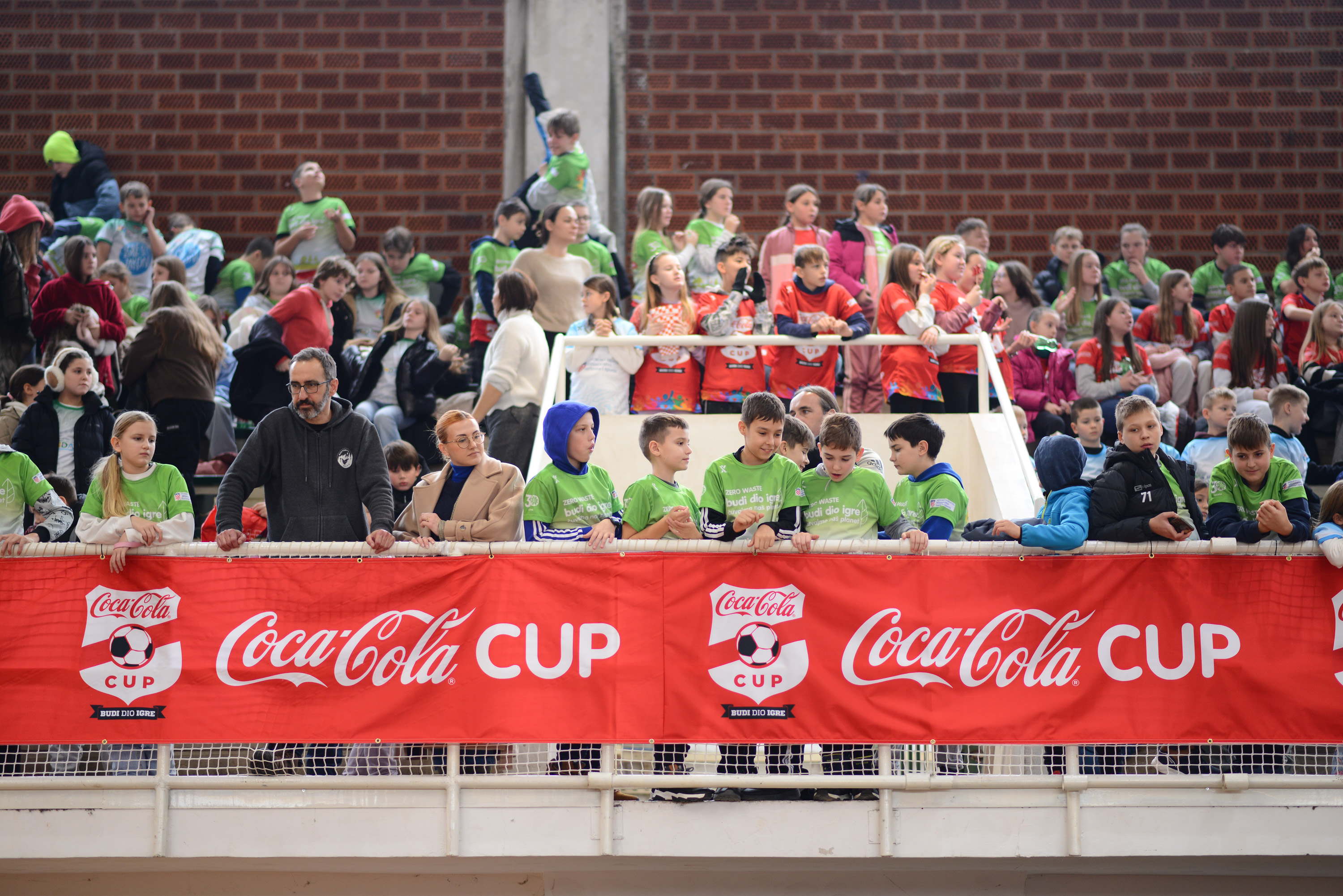 Djeca i mladi sudionici prate natjecanja s tribina tijekom Coca‑Cola Cupa, uz istaknute natpise turnira i sportsku atmosferu u dvorani.