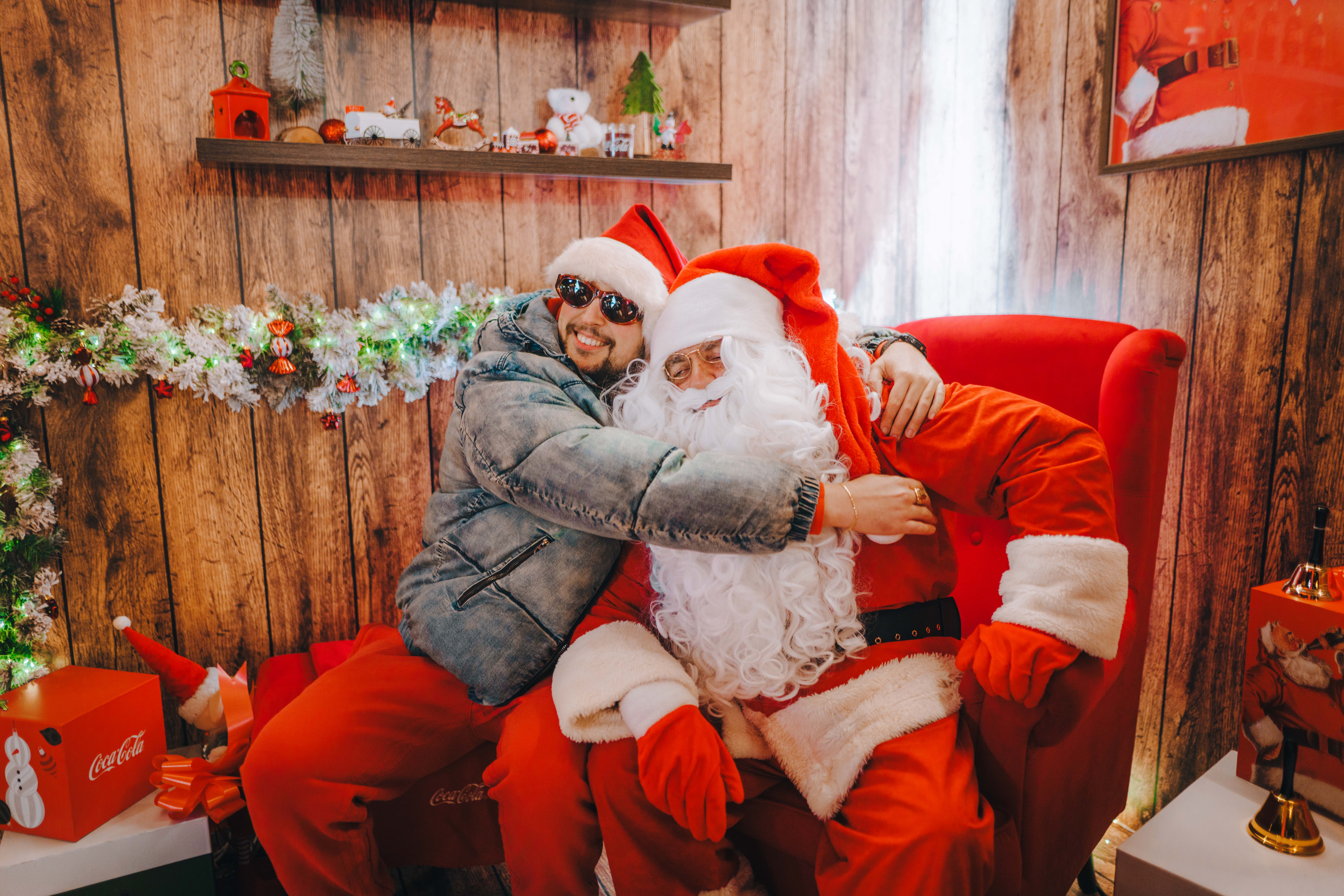 Posjetitelj grli Djeda Božićnjaka u blagdanski uređenom prostoru s božićnim dekoracijama, tijekom adventskog događanja Coca‑Cola