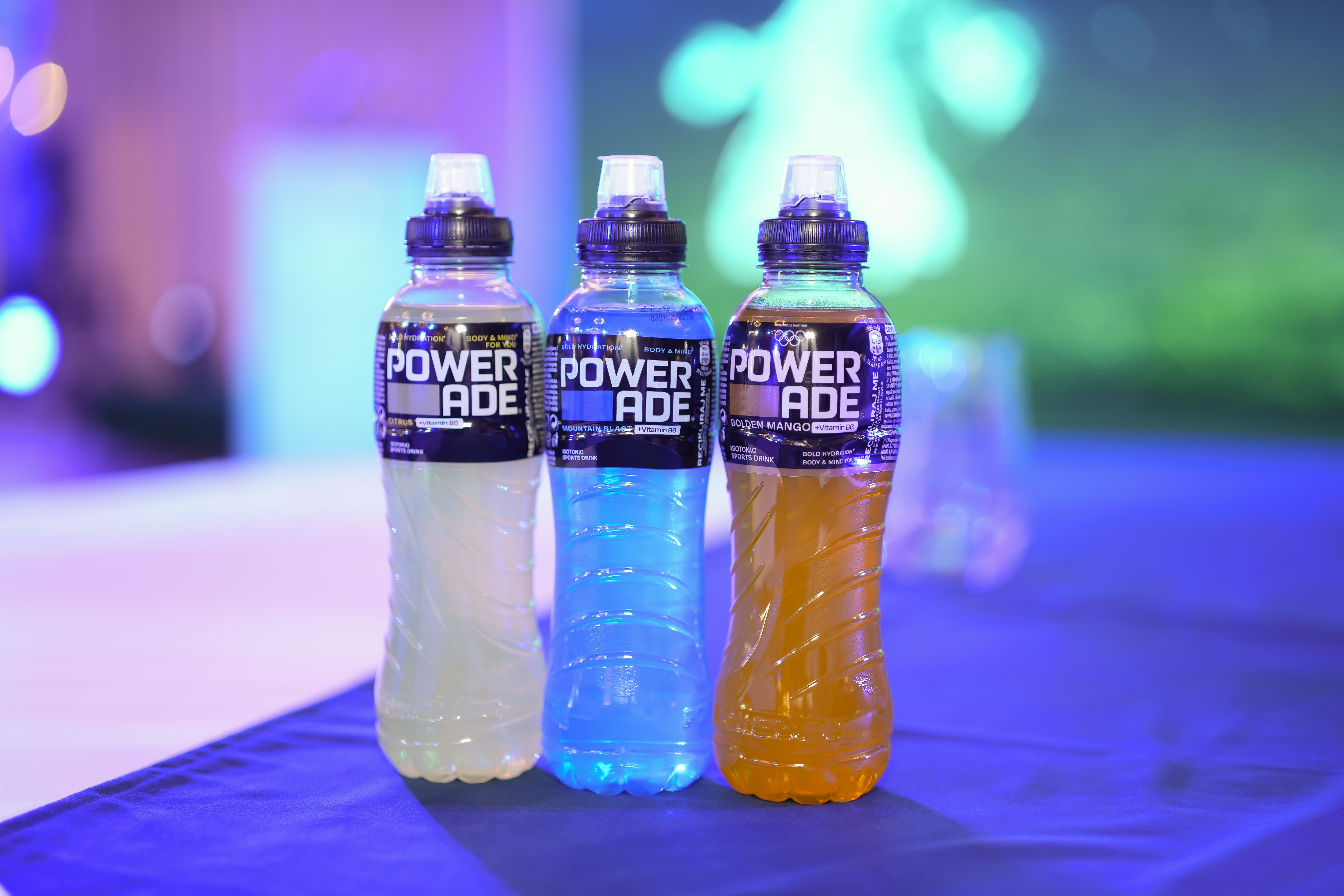 Tri boce Powerade sportskog napitka različitih okusa postavljene na stolu, s osvijetljenom pozadinom tijekom sportskog događaja