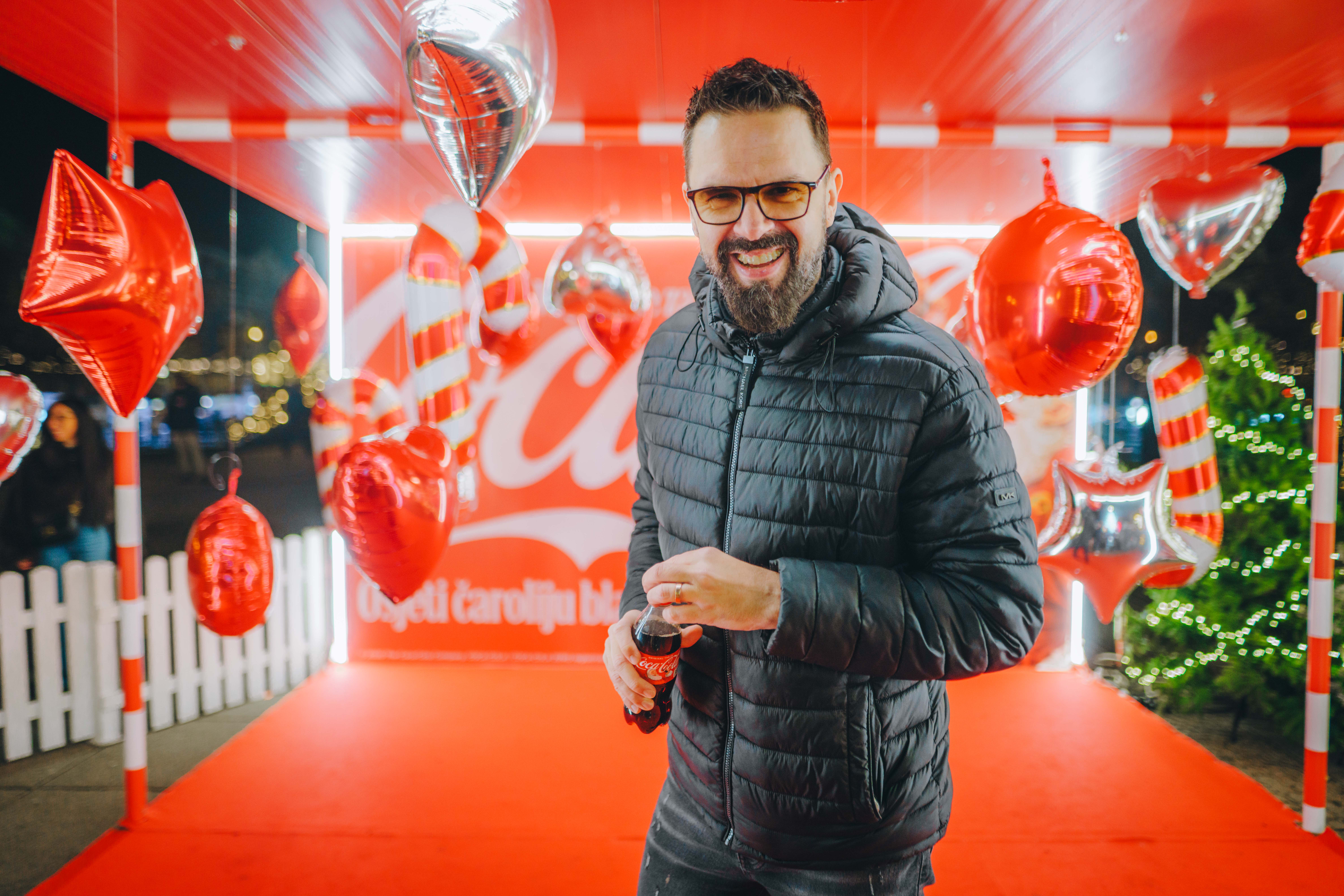 Posjetitelj stoji ispred Coca‑Cola božićnog kamiona okružen crvenim blagdanskim dekoracijama i drži čašu pića tijekom adventskog događanja.