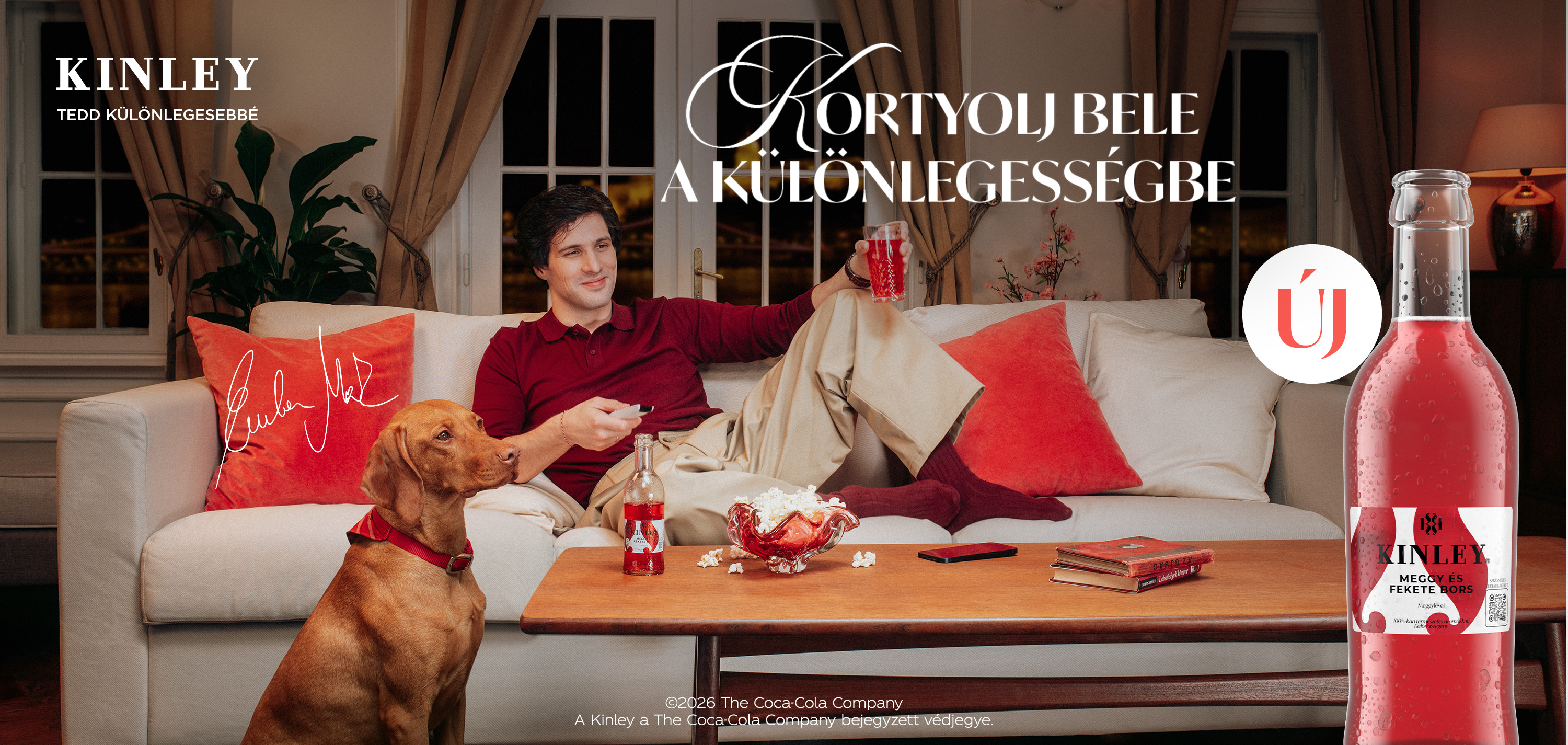 Kanapén ülő férfi Kinley itallal, kutya figyel, modern nappali, párnák és dohányzóasztal.