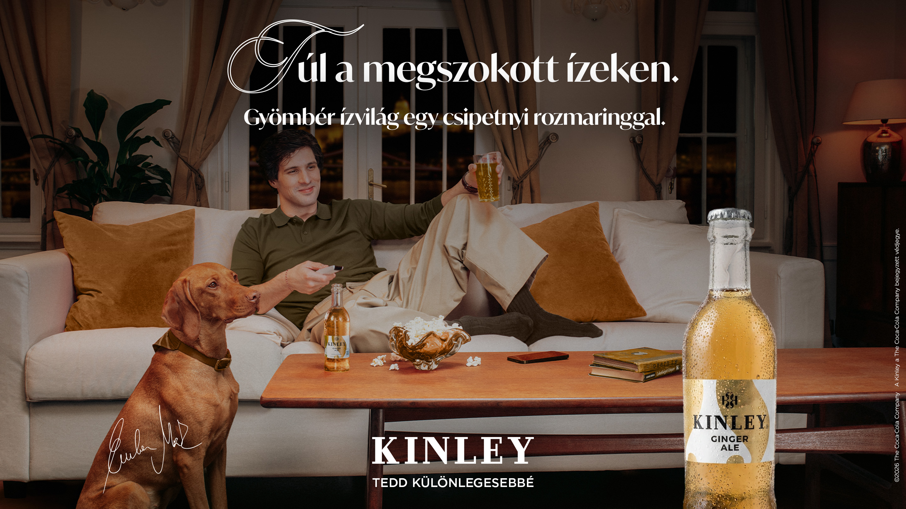 Kanapén ülő férfi Kinley itallal, kutya figyel, modern nappali, párnák és dohányzóasztal.