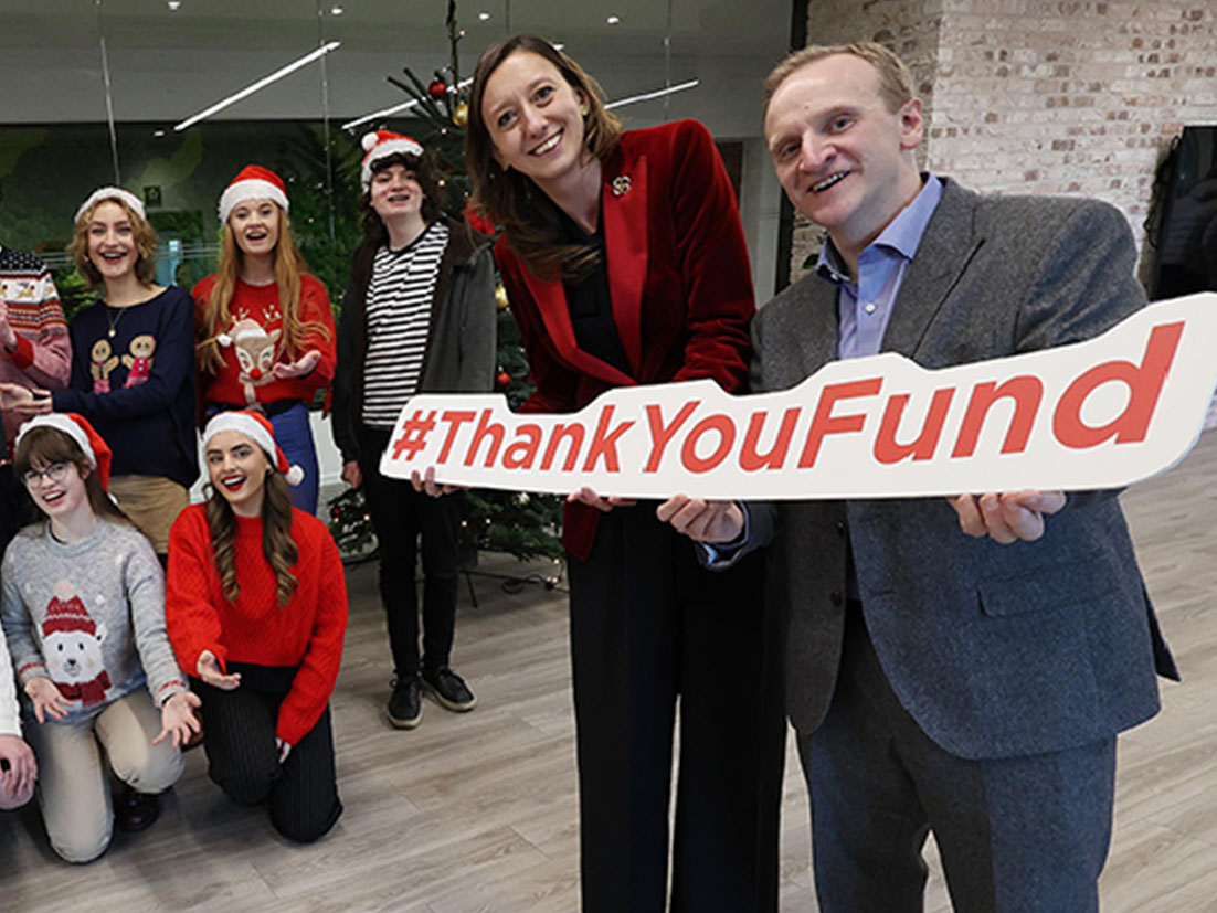 Group of people wearing christmas hats holding a Coca-Cola Thank You Fund certification and two people in suits on the side holding a Thank You Fund sign