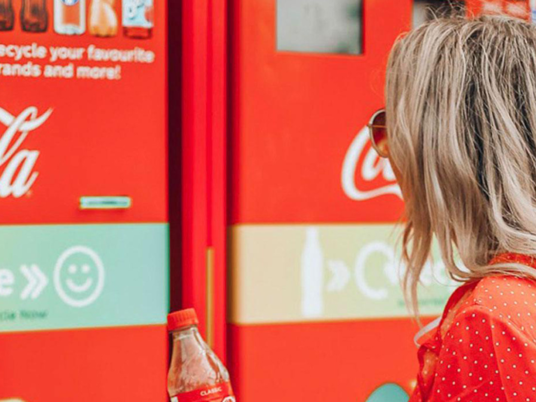 A woman holding an empty Coca-Cola bottle in front of a Coca-Cola themed recycle bin