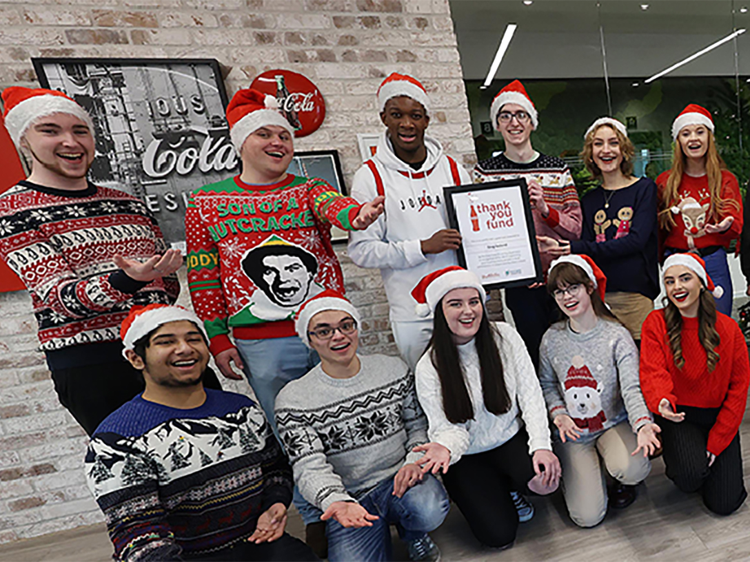 Group of people wearing christmas hats holding a Coca-Cola Thank You Fund certification and two people in suits on the side holding a Thank You Fund sign