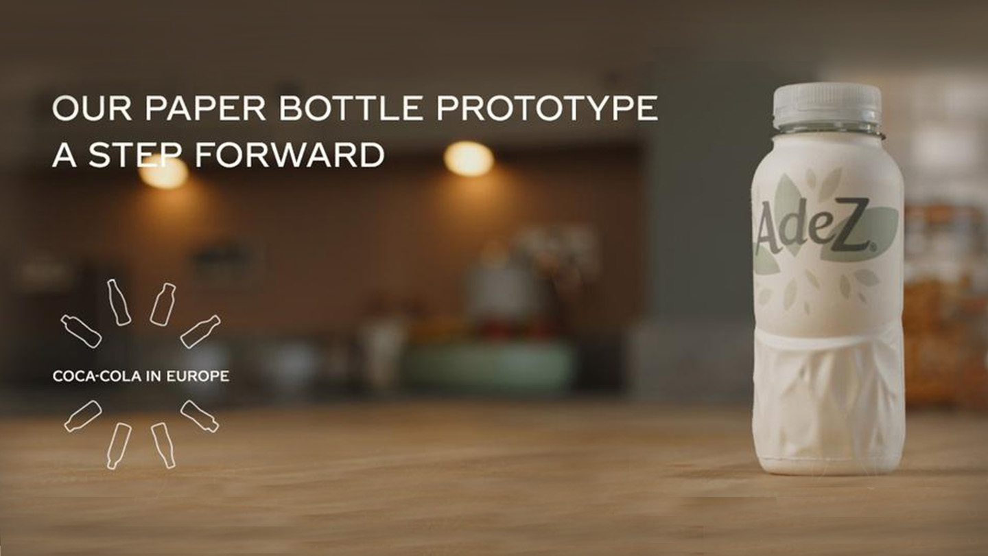 Coca-Cola paper bottle in the foreground with an out-of-focus background and the phrase 'Our paper bottle prototype. A step forward' displayed on the top of the image