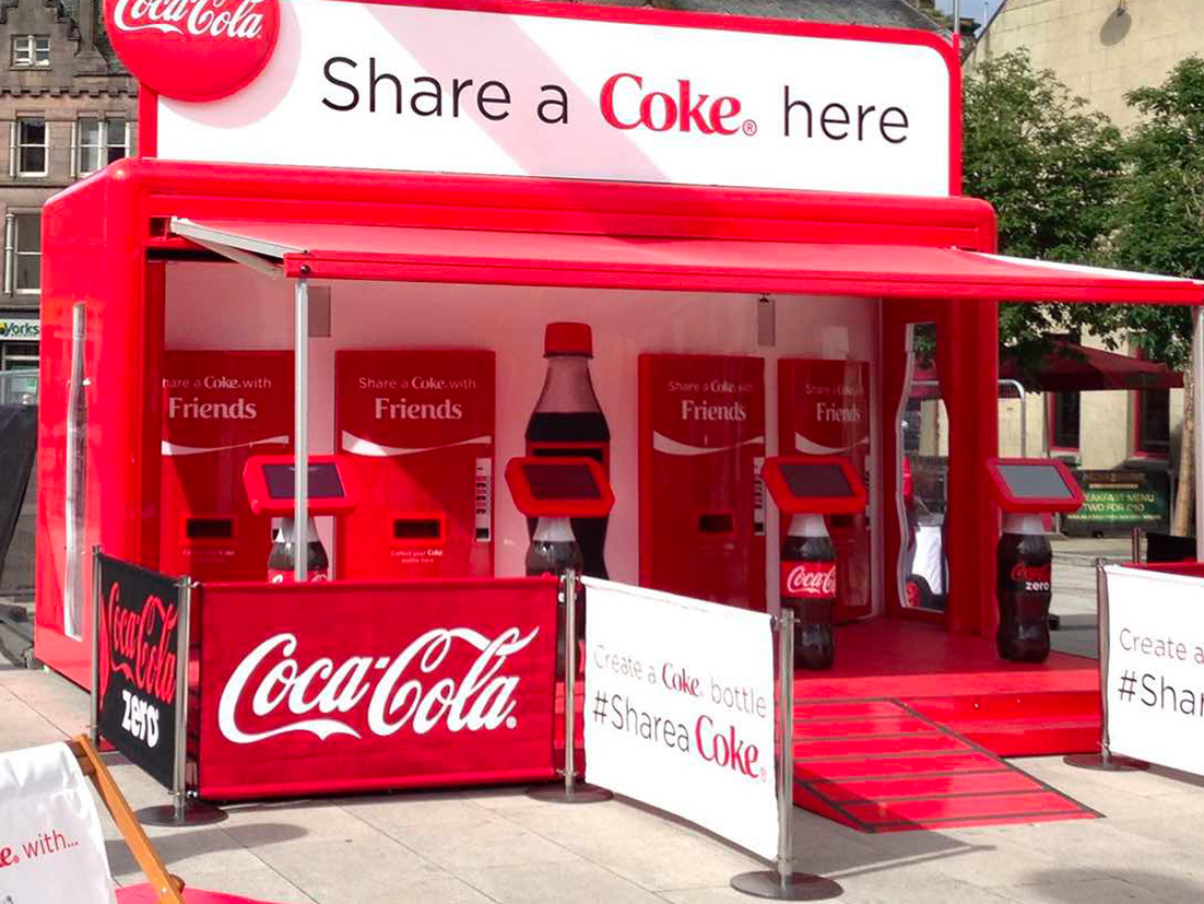 Coca-Cola structure in a city plaza with a banner with the phrase 'Share a Coke here' placed on top of it