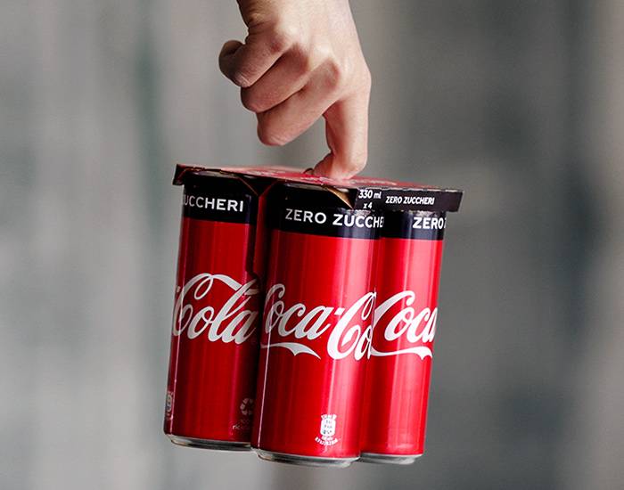 Mano che tiene quattro lattine di Coca-Cola Zero Zuccheri con nuovo packaging in cartone.