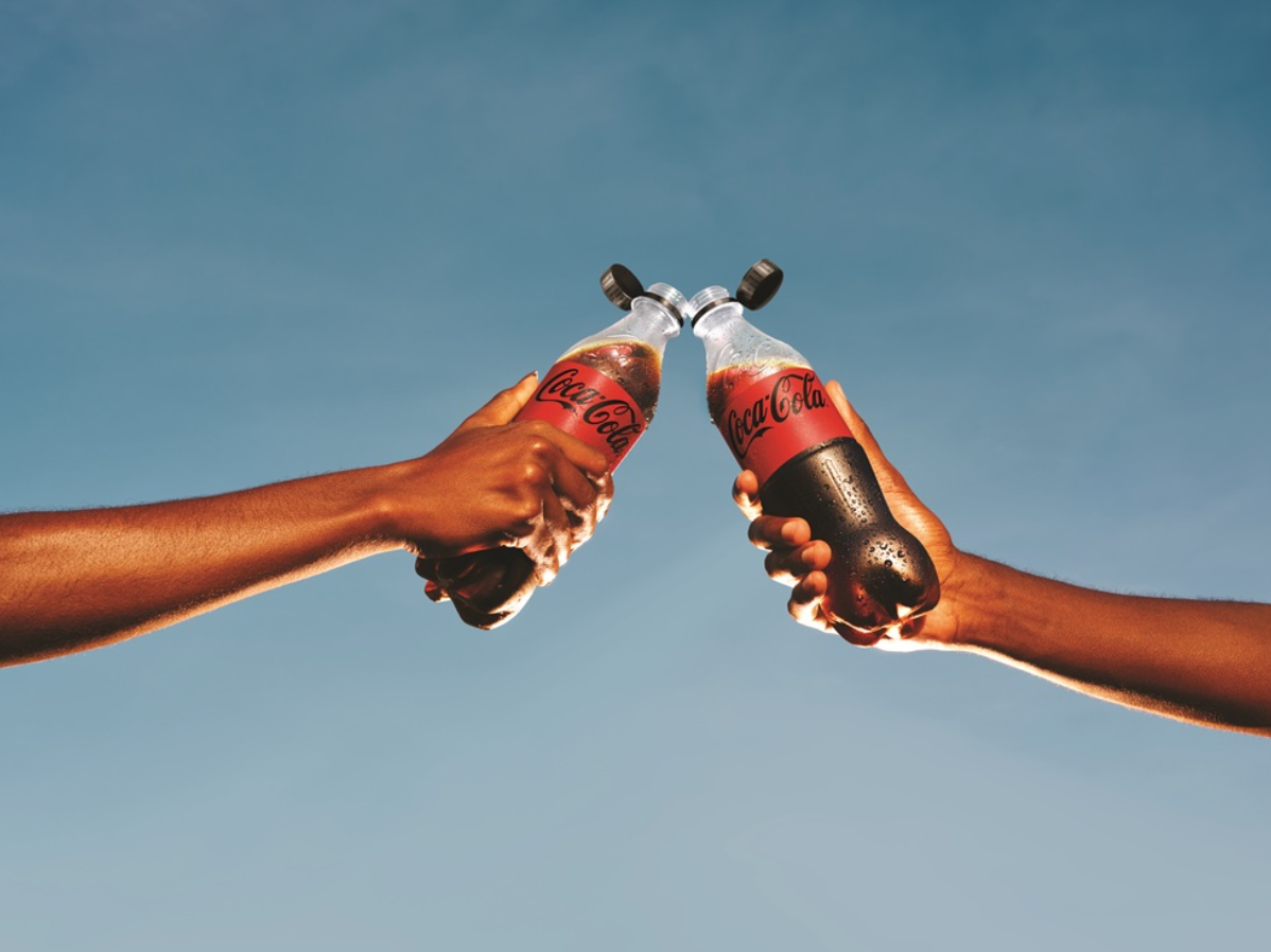 Due mani che reggono bottiglie di Coca-Cola con un cielo azzurro sullo sfondo.